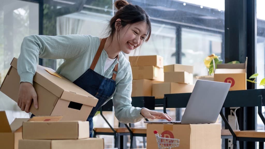 A shop owner checking orders on a laptop beside packed boxes